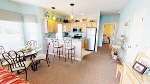 A compact kitchen with a central island, bar stools, and a dining area with a table and chairs. - Escape to River Creek | The Perfect NC Golf Condo | RC 1602 (Sunset Beach)