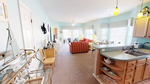 A spacious living area with a sofa, chairs, a glass coffee table, a kitchen with a sink and cabinets, and a dining area with a table and chairs. - Escape to River Creek | The Perfect NC Golf Condo | RC 1602 (Sunset Beach)