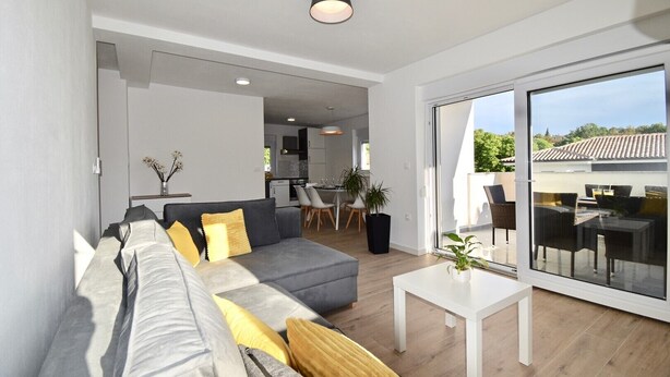 En moderne stue med en grå sofa, et lille hvidt bord og en glasdør, der fører til et udendørs siddeområde.