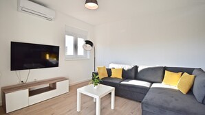 En moderne stue med et fladskærms-tv, en grå sofa med gule puder, et hvidt sofabord og en gulvlampe.