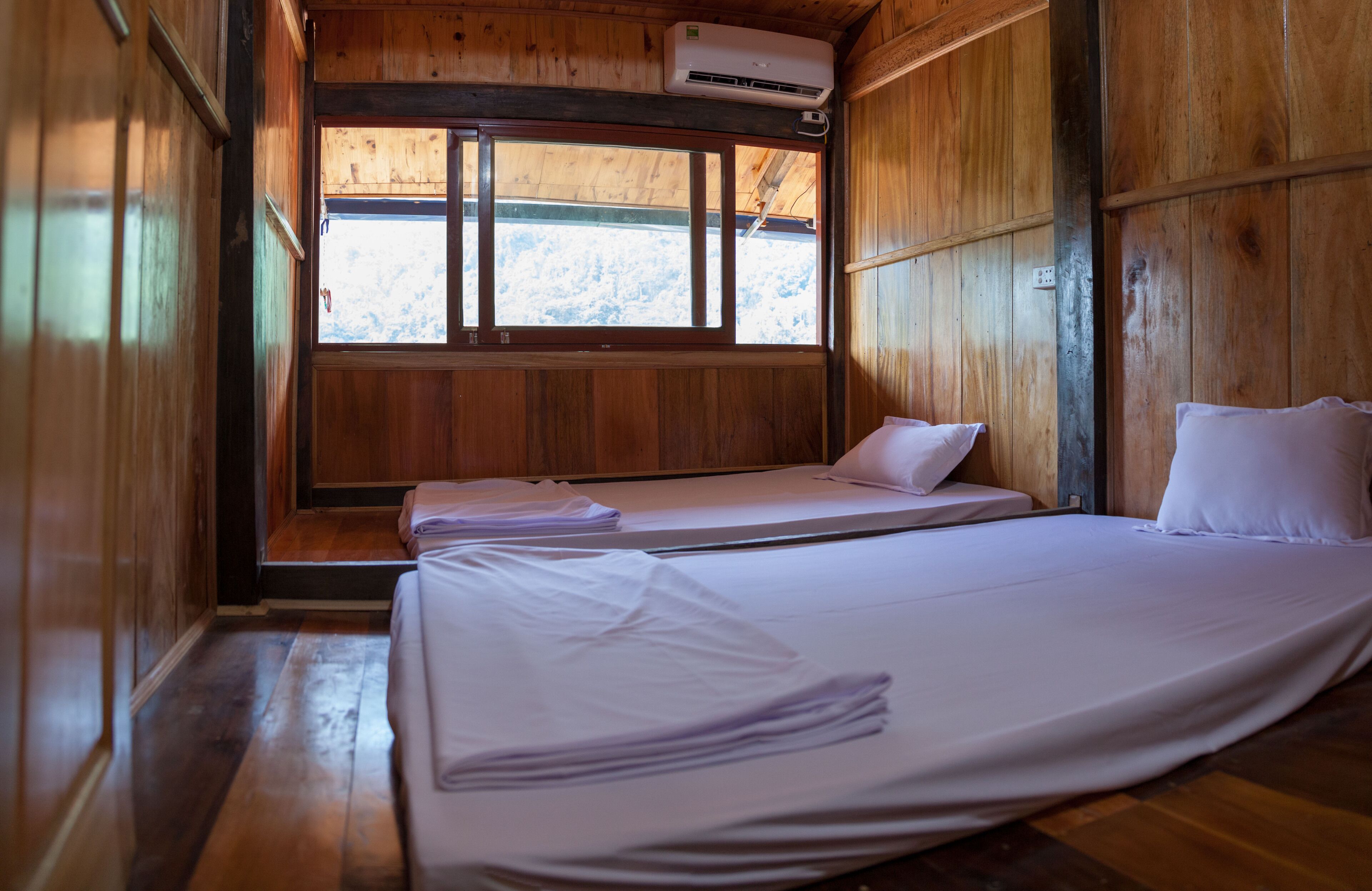 A wooden cabin room with two beds, a window, and an air conditioning unit.