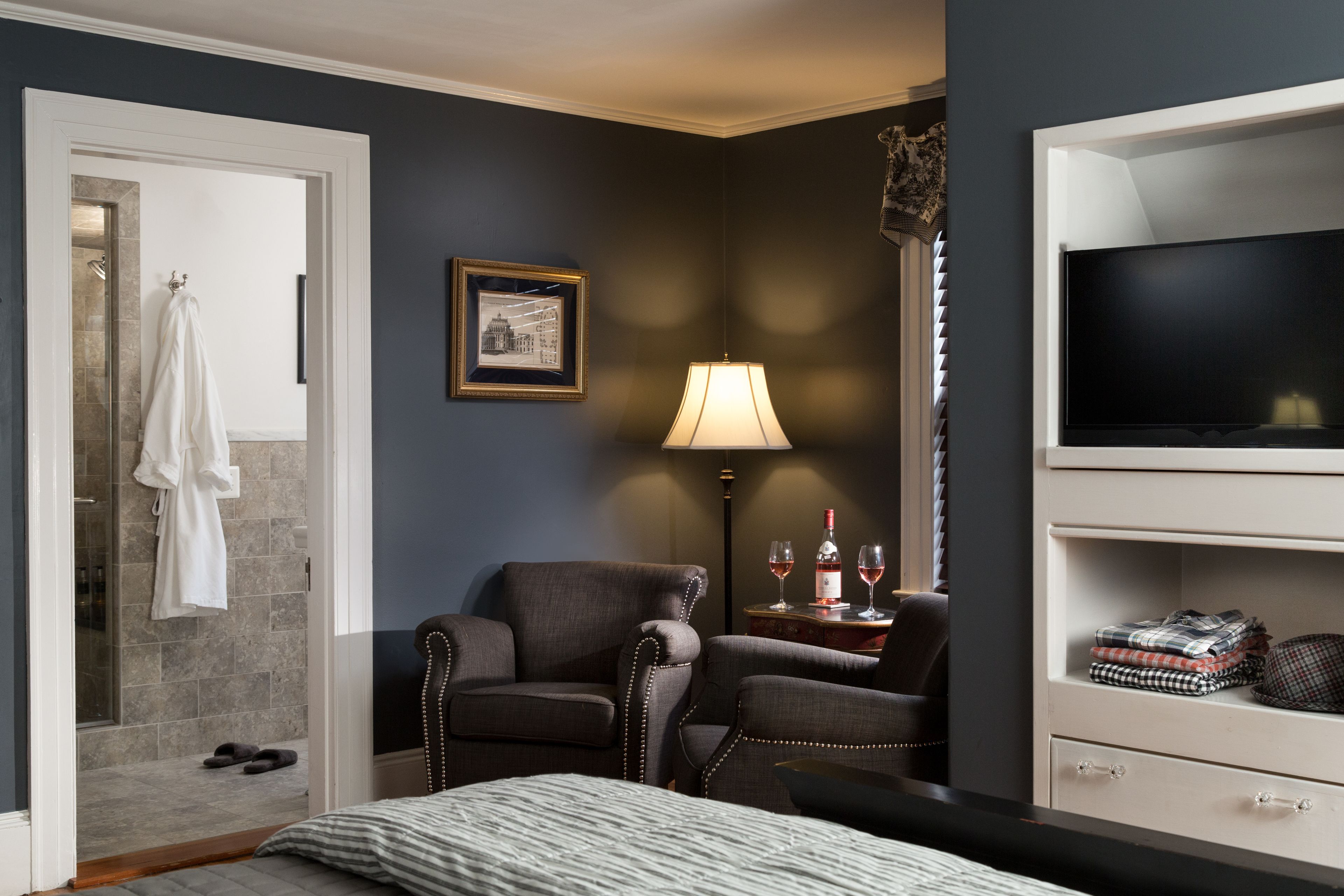 A hotel room with a bed, two armchairs, a TV, a lamp, a framed picture, and a small table with glasses and a bottle.