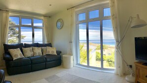 A living room with a dark leather sofa, a round clock, and a large window offering a view of the sea and buildings.
