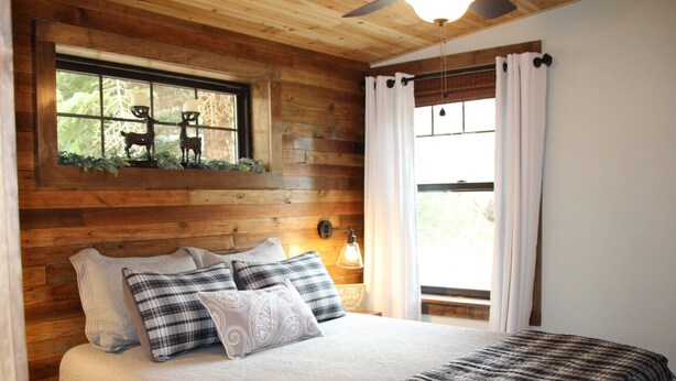 A bedroom with wooden walls, a bed with plaid bedding, a window with a view of trees, and a wall-mounted light fixture. - Great Getaway on the Big Thompson very near Estes & Rocky Mt. Nat'l Park (Drake)