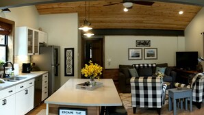 A modern kitchen with white cabinets, a central island, and a dining area with checkered chairs. There is a television and framed pictures on the wall. - Great Getaway on the Big Thompson very near Estes & Rocky Mt. Nat'l Park (Drake)