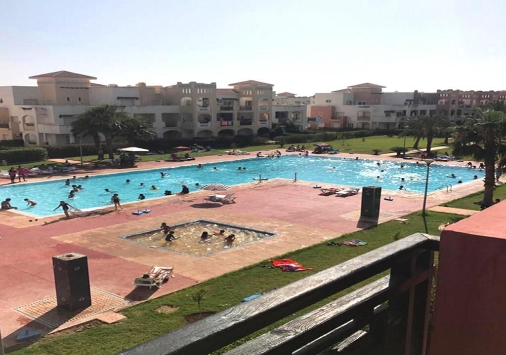 A swimming pool area with multiple pools, lounge chairs, and a building complex in the background.