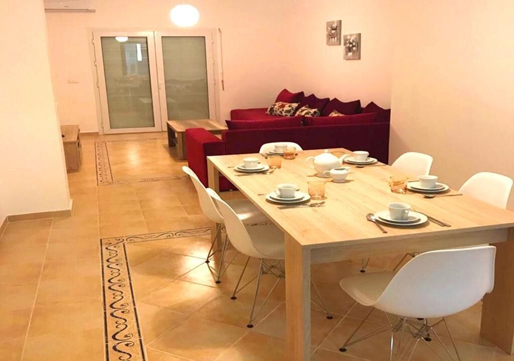 Un coin repas avec une table en bois dressée pour quatre personnes, des chaises blanches, un canapé rouge et une porte-fenêtre coulissante donnant sur un balcon.