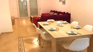 Un comedor con una mesa de madera dispuesta para cuatro, sillas blancas, un sofá rojo y un gran ventanal corredizo que da a un balcón.