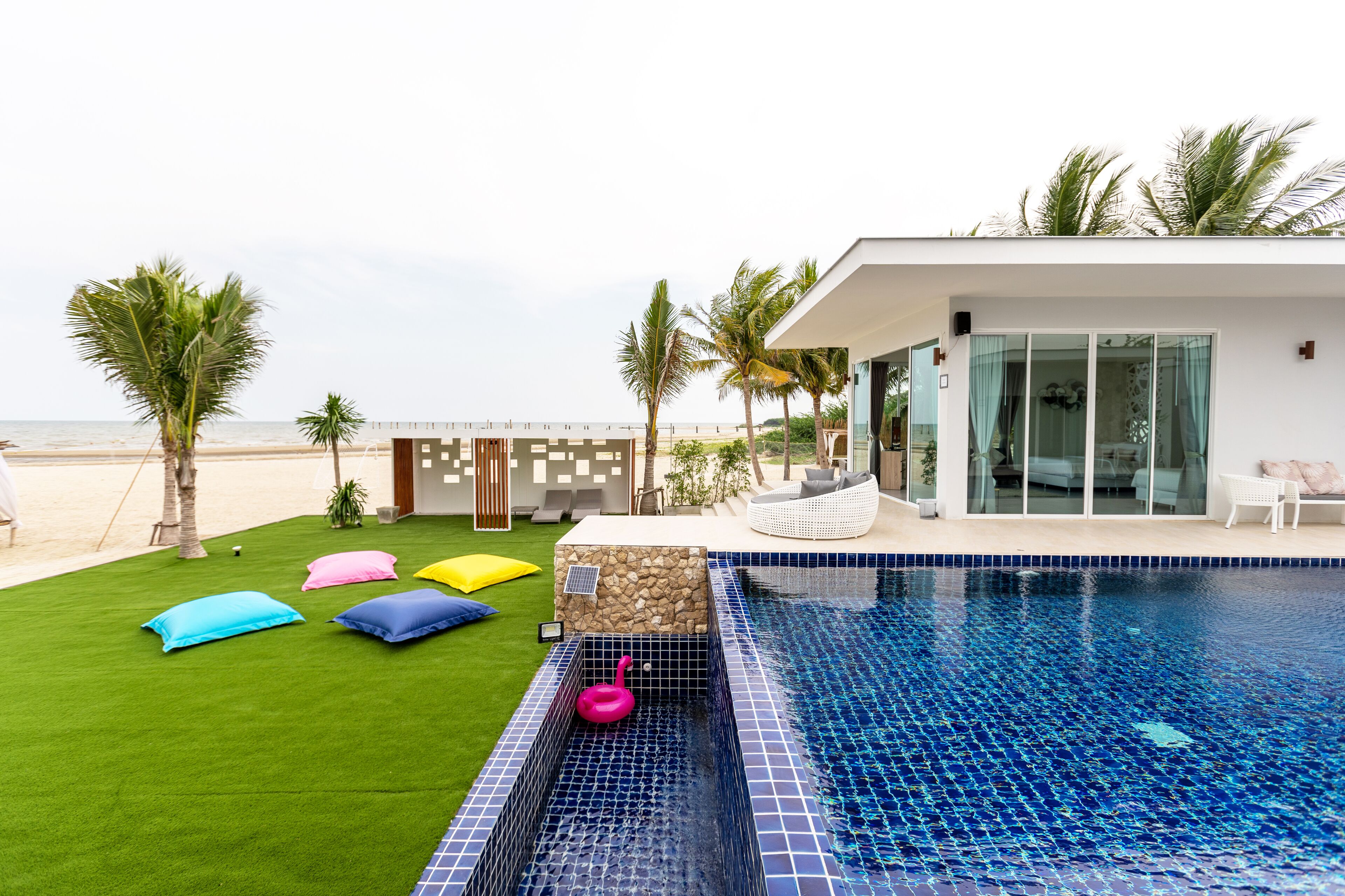 Ein moderner Poolbereich mit freiem Blick auf den Ozean, eine Rasenfläche mit bunten Kissen und ein zeitgemäßes Gebäude mit großen Fenstern.