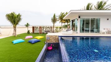 Ein moderner Poolbereich mit freiem Blick auf den Ozean, eine Rasenfläche mit bunten Kissen und ein zeitgemäßes Gebäude mit großen Fenstern.