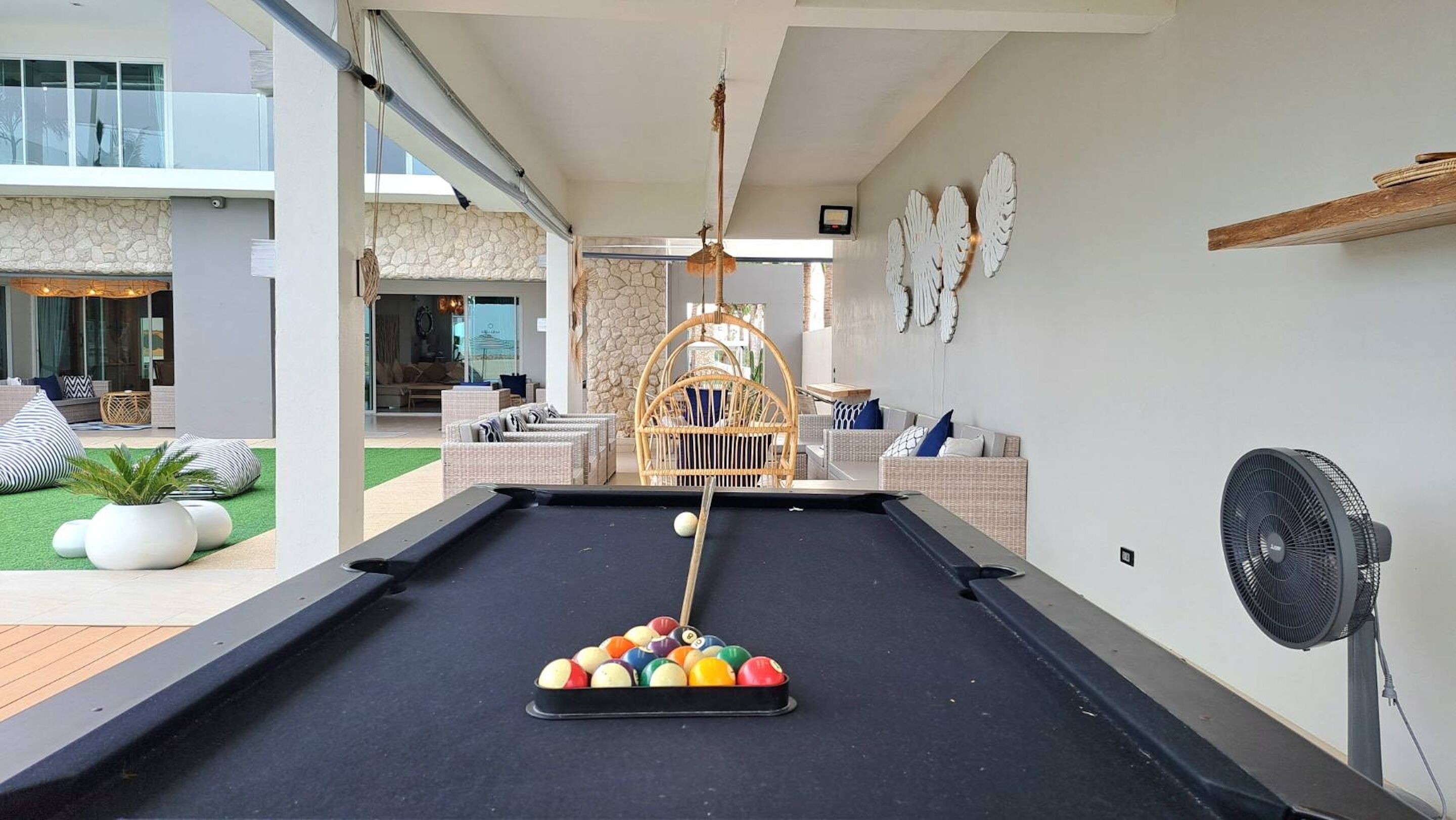 A pool table with a variety of balls, a fan, and a rattan chair in a modern indoor setting.