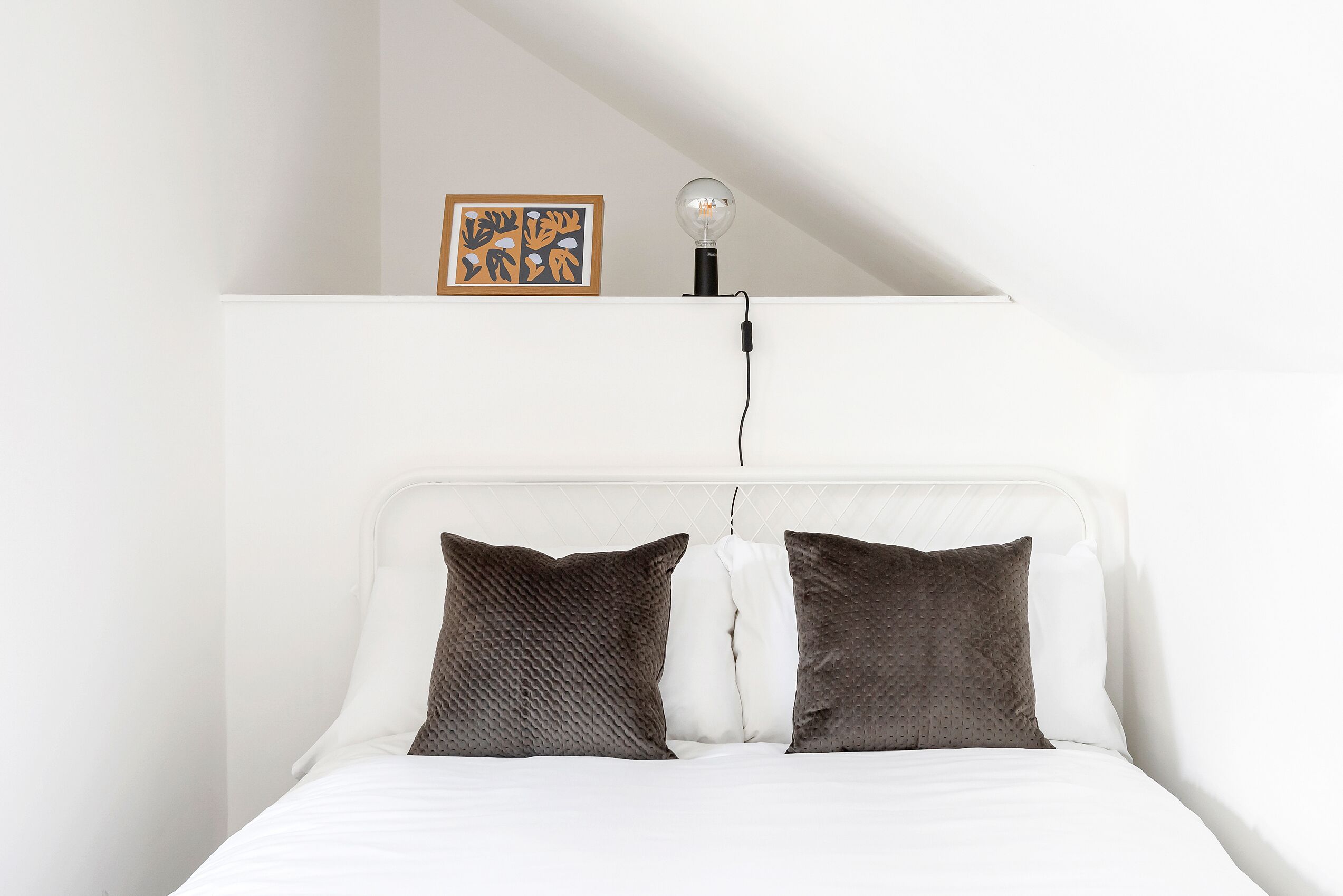 A neatly made bed with white and dark brown pillows, a single light bulb on a shelf above, and a framed artwork on the wall.