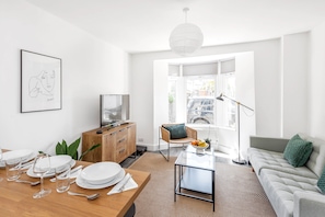 A modern living room with a dining area, a sofa, a coffee table, and a view of the street through large windows.