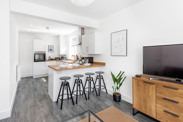 A modern kitchen with white cabinets, a wooden island with bar stools, a large flat-screen TV, and a potted plant.