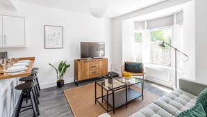 A modern living room with a sofa, a coffee table, a TV, and a dining area with stools and a table set for a meal. - Stylish Apartment In Leafy Central Neighbourhood (Cardiff)