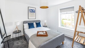 A modern hotel room with a neatly made bed, a bedside table with a lamp, a window with a view, and a hanging rack. - Stylish Apartment In Leafy Central Neighbourhood (Cardiff)