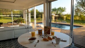 A breakfast setup with a table, breakfast items, and a view of a swimming pool and garden.