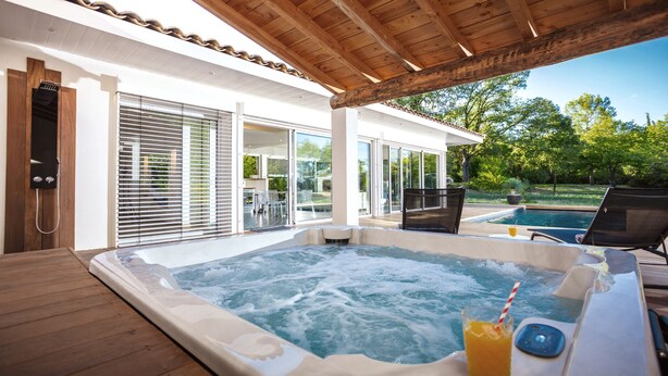 A hot tub area with a wooden deck, a glass-enclosed patio, and a view of a swimming pool and greenery. - Villa with private pool and spa (CARNAS)