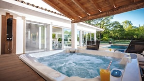 A hot tub area with a wooden deck, a glass-enclosed patio, and a view of a swimming pool and greenery.