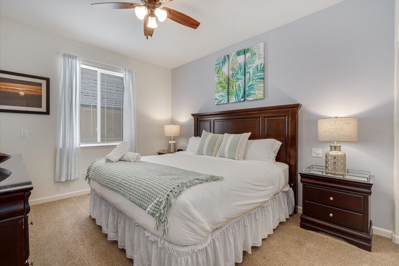 Une chambre à coucher avec un grand lit, un ventilateur de plafond, une table de chevet avec une lampe et une fenêtre avec des rideaux.
