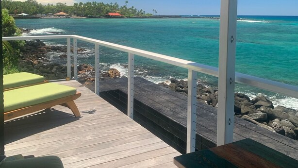 Une terrasse en bois avec deux chaises vertes à coussins et une table en bois, d’où l’on peut admirer une mer bleue et limpide, une côte rocheuse et des arbres à l’horizon.