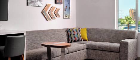 A modern living room with a grey sectional sofa, a small round table, and colorful cushions. There are abstract wall art pieces and a large window with a city view.