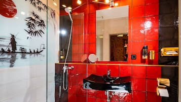 A modern bathroom with a red and black tiled wall, a glass shower enclosure, and a black sink with a gold faucet.