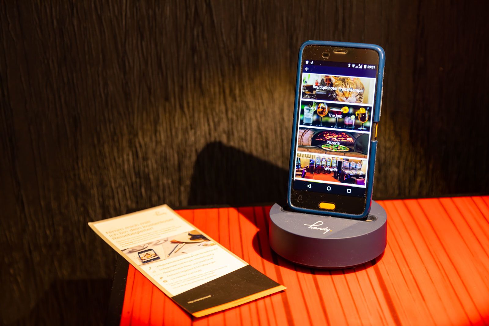 A smartphone on a stand displaying a hotel app with various sections like 'Boutiquehotel', 'The jam', 'Pizzeria', and 'Winwin'.