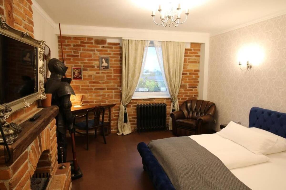 A hotel room with a brick wall, a bed with a blue headboard, a desk with a chair, a radiator, and a chandelier.