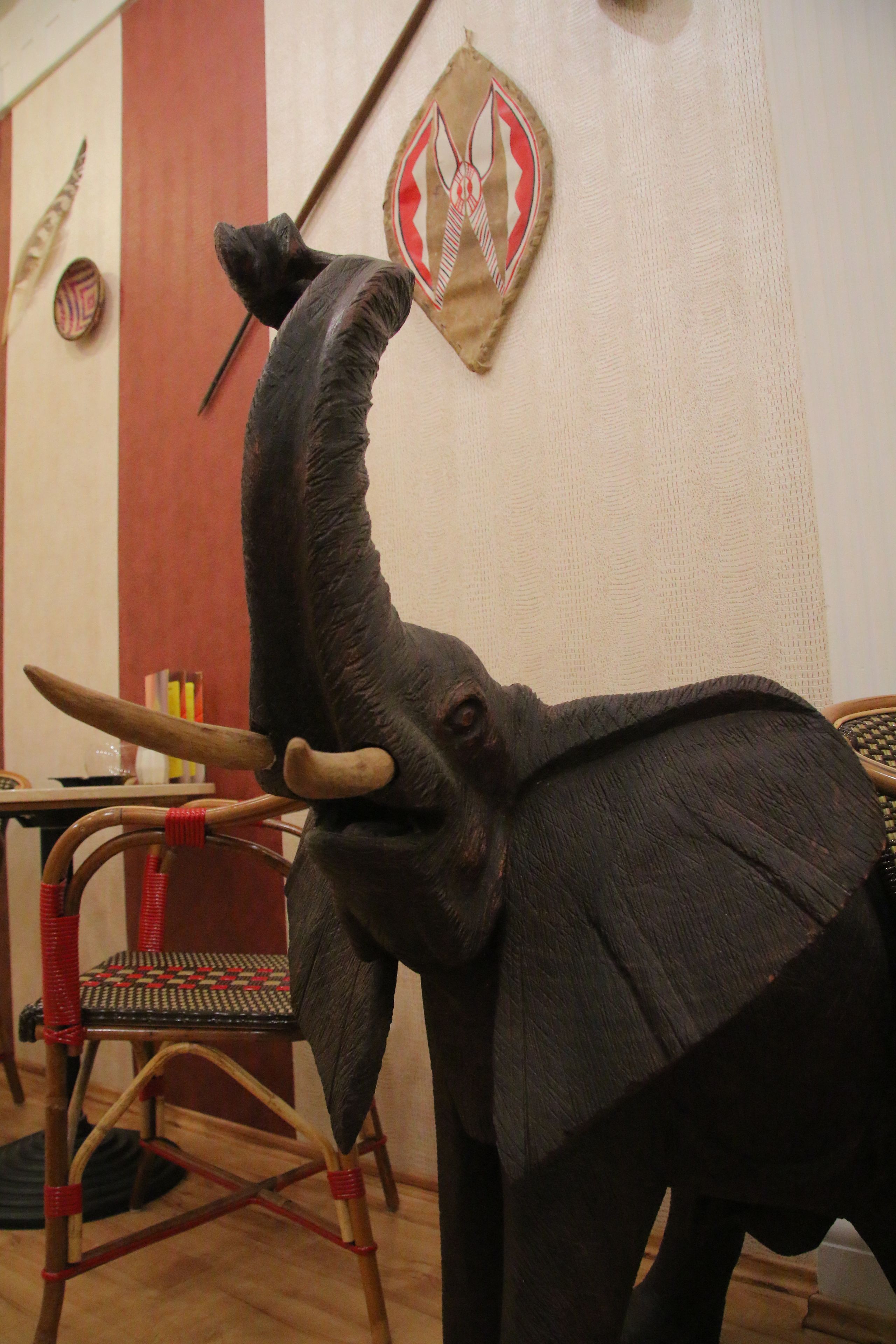 Una habitación con una escultura decorativa de cabeza de elefante, una silla tejida y un escudo en la pared.