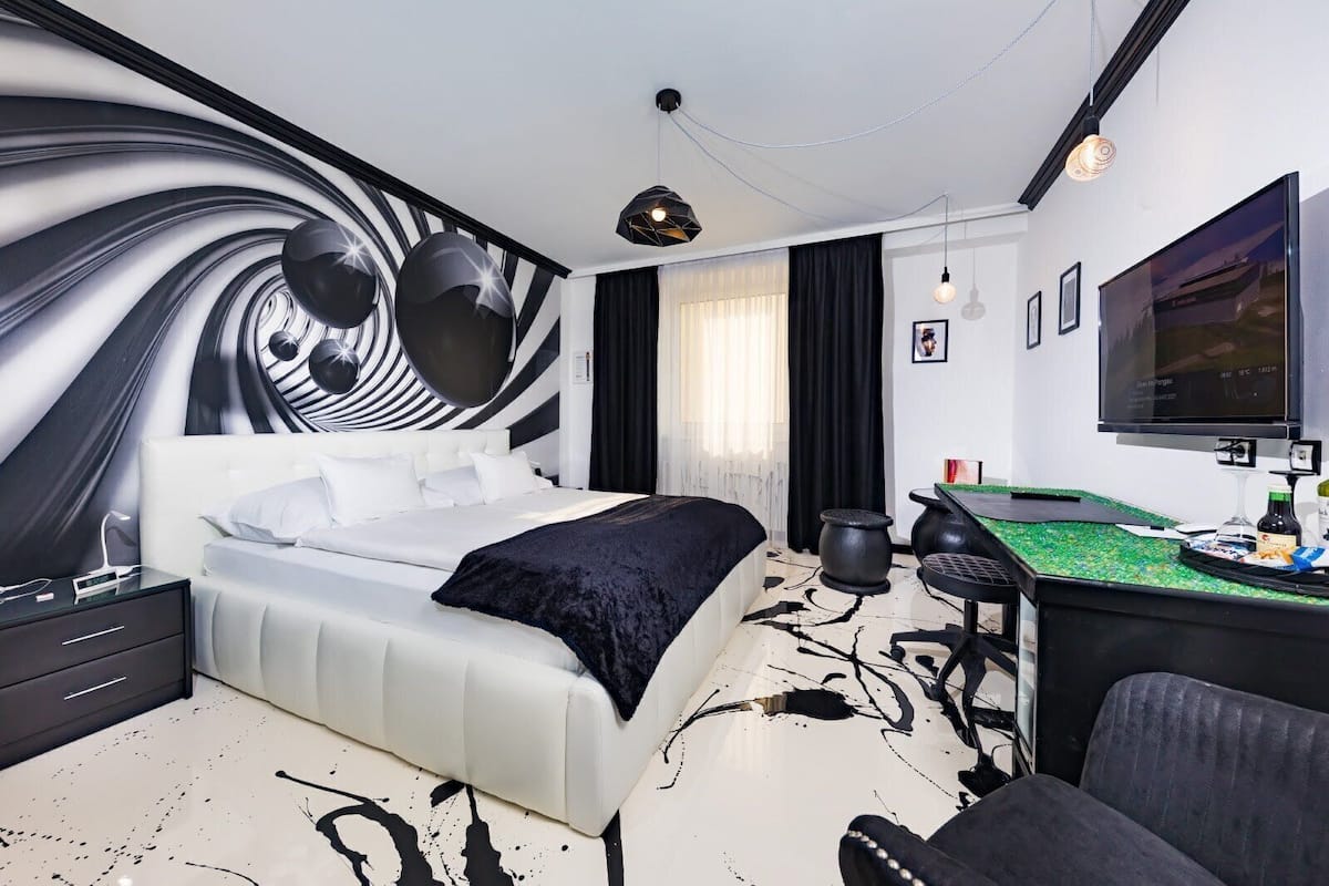 A modern hotel room with a large bed, a desk with a green table, a flat-screen TV, and a unique black and white wall mural.