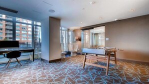 A hotel room with a large window, a desk with a chair, a foosball table, and a patterned carpet. - Luxury 1BR | Health Club | Pool | Near PATH (Jersey City)