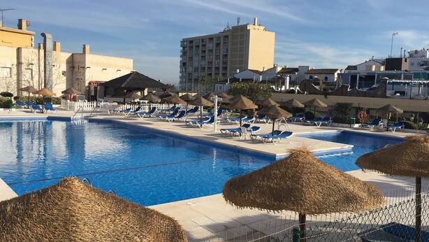Une piscine entourée de bâtiments, agrémentée de chaises longues et de parasols en chaume.