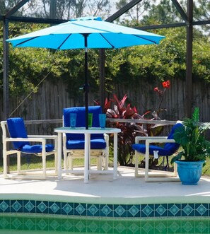 An outdoor patio with a blue umbrella, white chairs, a table, and a potted plant.