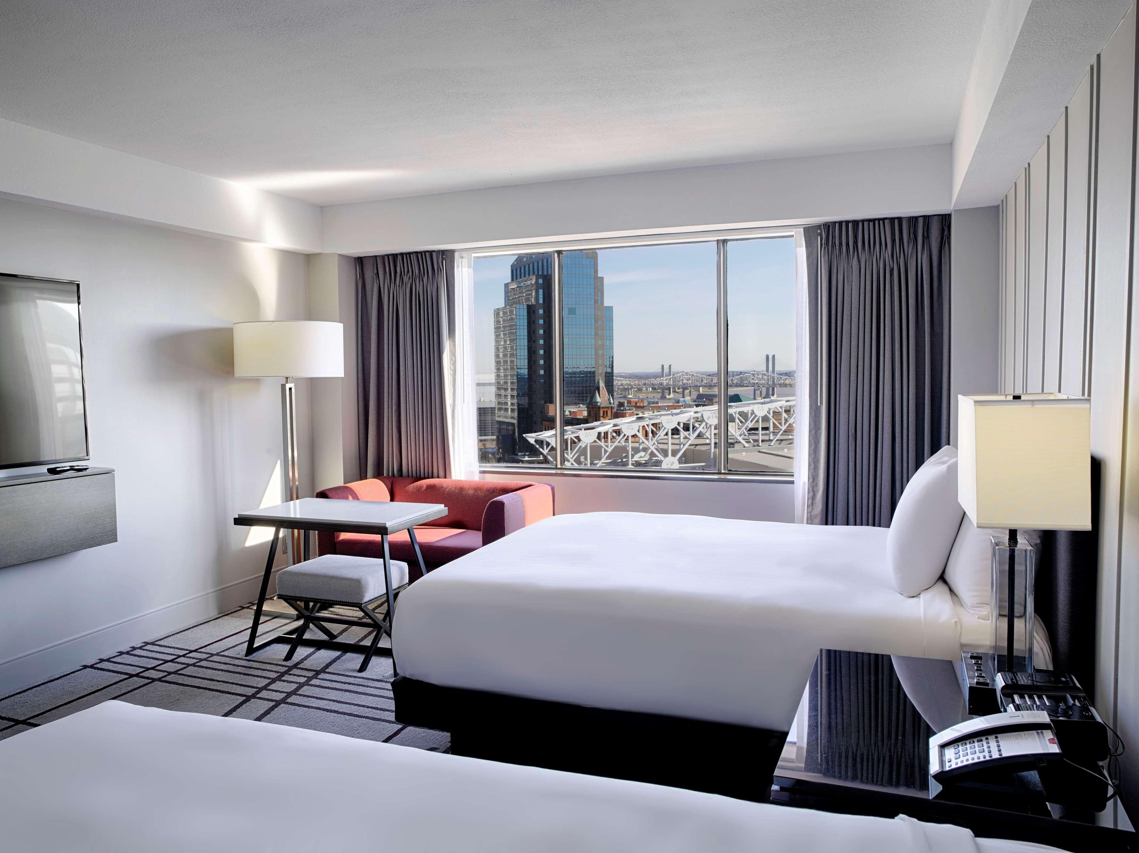 Ein Hotelzimmer mit Bett, Schreibtisch, Sessel, Fernseher und Blick auf eine Stadtlandschaft.