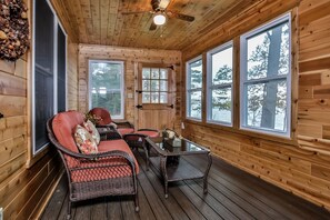 Una cabaña de madera con una zona de estar acogedora, que incluye sillones de mimbre y una mesa, ventanas amplias que ofrecen vistas a los árboles y un ventilador de techo.
