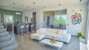 Un salon moderne avec un canapé blanc, une table basse agrémentée d’un arrangement floral, un coin cuisine avec un bar et un coin repas offrant une vue agréable.