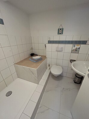 Et badeværelse med bruser, toilet, vask og en lille hylde med håndklæder.