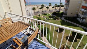 Ein Balkon mit Holztisch und -stühlen, der Blick auf einen Strand mit Palmen und einem wolkenlosen Himmel bietet.