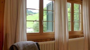 A room with a view of a green landscape through a window with a wooden frame and white curtains. - Chalet Alte Post by Interhome (Schwenden)