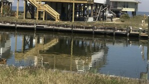 Une maison à deux étages avec un toit bleu, une terrasse en bois et un quai qui s’avance sur une étendue d’eau.