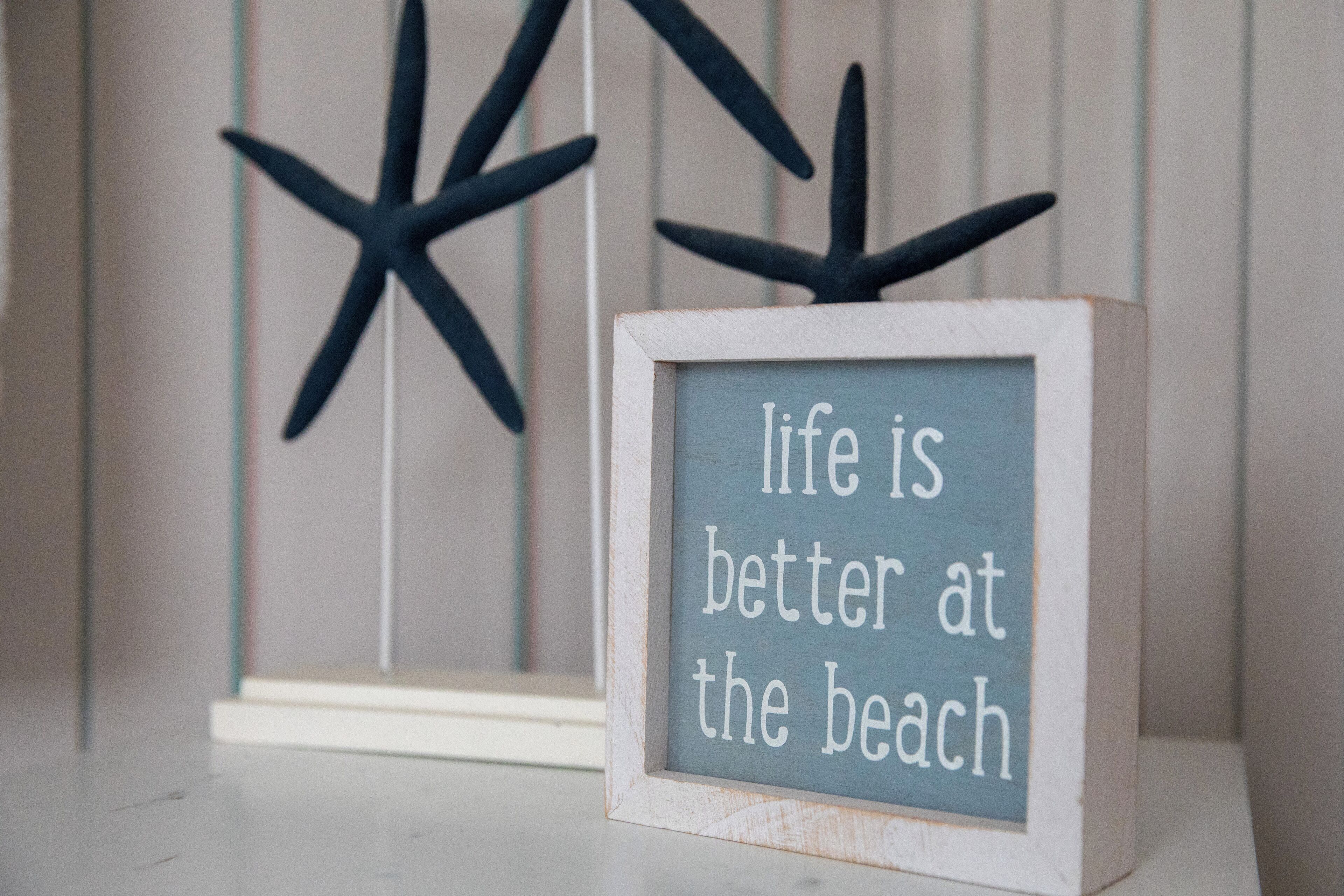 Une affiche encadrée avec l’inscription « la vie est plus belle à la plage », posée sur une étagère.
