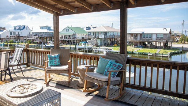 Une terrasse en bois, agrémentée de chaises et d’une table en osier, qui offre une vue sur une marina avec des bateaux et des maisons.