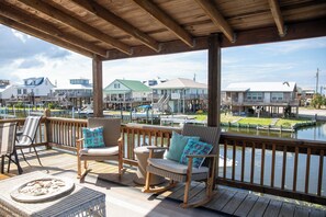 A wooden deck with wicker chairs and a table, overlooking a marina with boats and houses.
