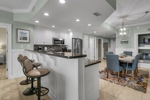 A modern kitchen with white cabinets, a granite countertop, and stainless steel appliances. There is a bar area with stools and a dining table with chairs.