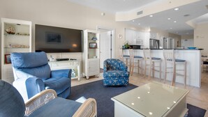 Een moderne woonkamer met een flatscreen televisie, een blauwe bank, een glazen salontafel, en een keuken met witte kasten en een barkruk.