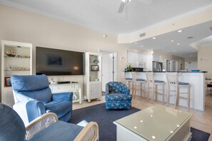 A modern living room with a flat-screen TV, a blue sofa, a glass coffee table, and a kitchen area with white cabinets and a bar stool.