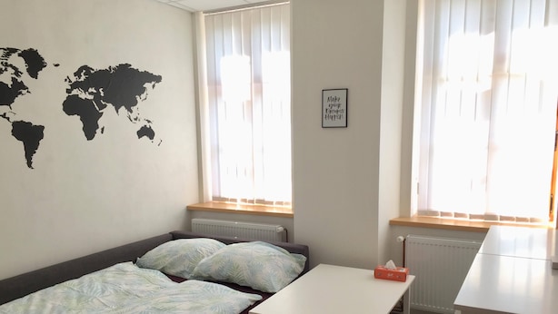 A small, well-lit room with a bed, a small table, and a world map on the wall.