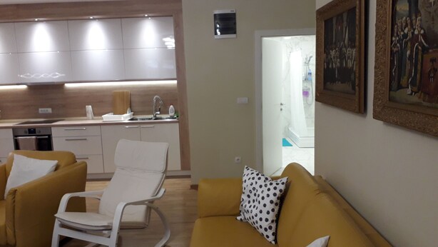 A modern living room with a yellow sofa, a white rocking chair, and a dining area with a kitchen in the background. - Silicon Valley Apartments (Stari Grad, Belgrade)