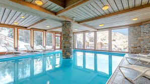 An indoor swimming pool with wooden beams and stone pillars, surrounded by lounge chairs and large windows offering mountain views. - Appartement avec balcon/terrasse | Accès piscine (Aime)